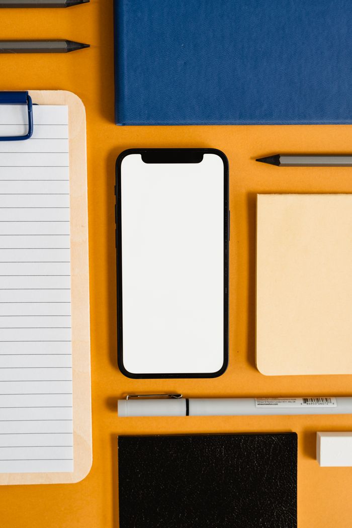 Flat lay of office stationery items with a blank smartphone screen on an orange background.